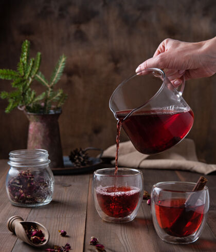 茶一只手把红玫瑰茶从一个玻璃茶壶倒进一个棕色木制的透明杯子里表.正面查看香料叶新鲜