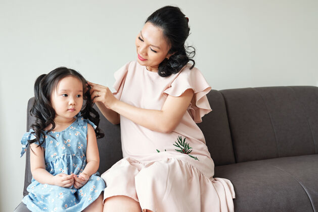 人一位怀孕的越南年轻女子在早上准备时给她严肃的小女儿编头发父母头发爱