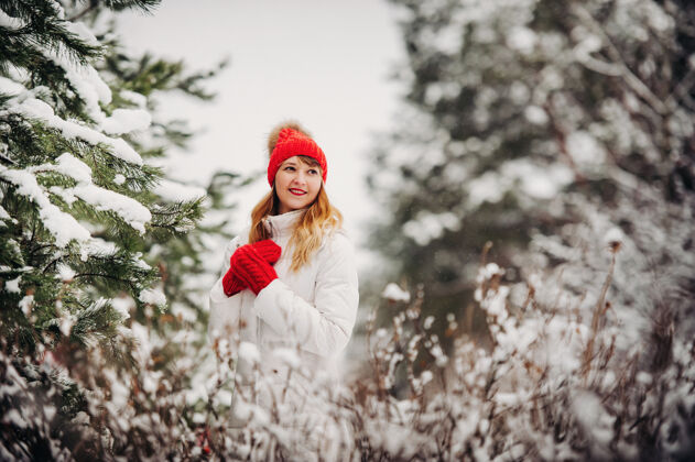框架一个穿着白衣服 戴着红帽子的女人在寒冷的冬天的肖像森林女孩在冰雪覆盖的冬季森林里女人霜冻微笑