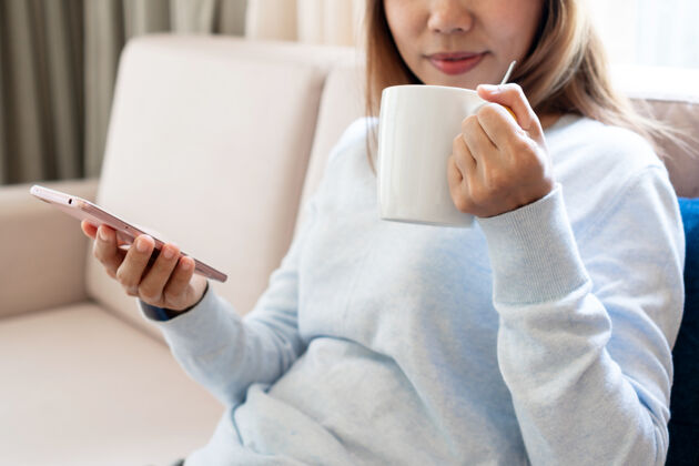 舒适亚洲女人放松 在家沙发上喝咖啡的时候用手机杯子科技女人