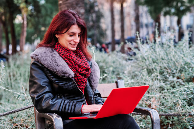 生活方式坐在公园长凳上使用笔记本电脑的女人户外科技城市理念无线人女人