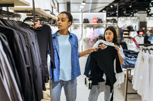 人两个女孩把衣服放在衣架上女店员在时尚精品店购物 购物狂 看服装的购物者女人零售选择