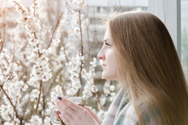 栗子拿着一杯茶的女孩站在窗前 望着窗外距离.开花背景中的树开花女人茶
