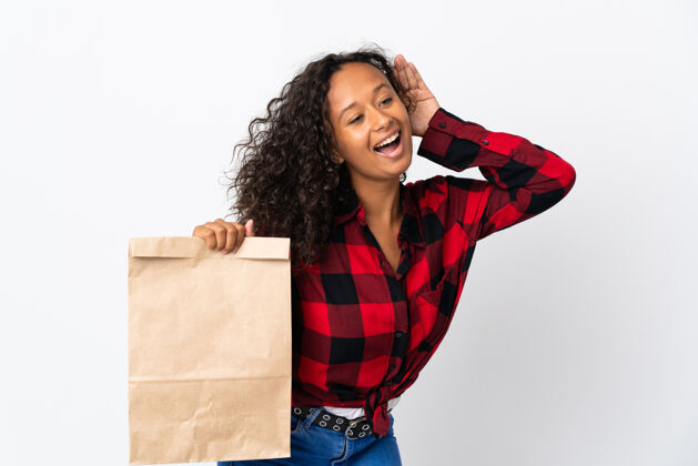 购物者一个十几岁的女孩拿着一个杂货店的购物袋去外卖 在白色的背景下听一些东西 手放在耳朵上消费者工作室人