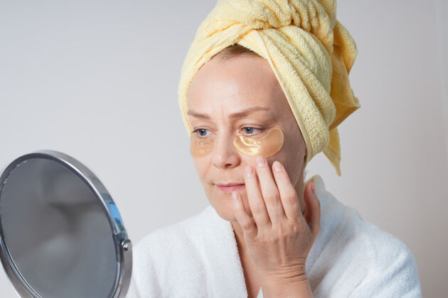 女人一个穿着白色晨衣的成熟女人 头上戴着一条黄色的毛巾 眼睛下面贴着眼罩 照镜子应用提升干净