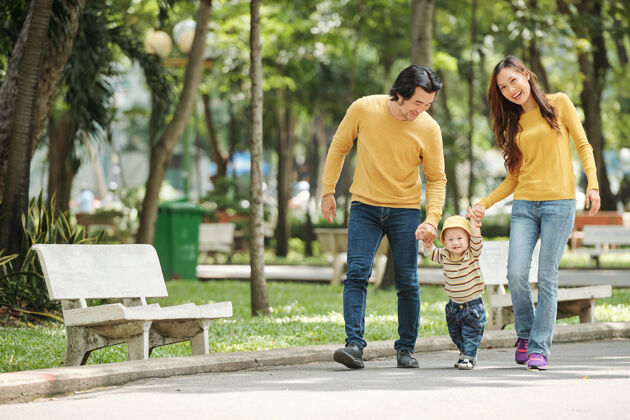 父母在阳光明媚的日子里 快乐的越南丈夫 妻子和穿着牛仔裤和毛衣的男婴在当地公园散步爱阳光成人