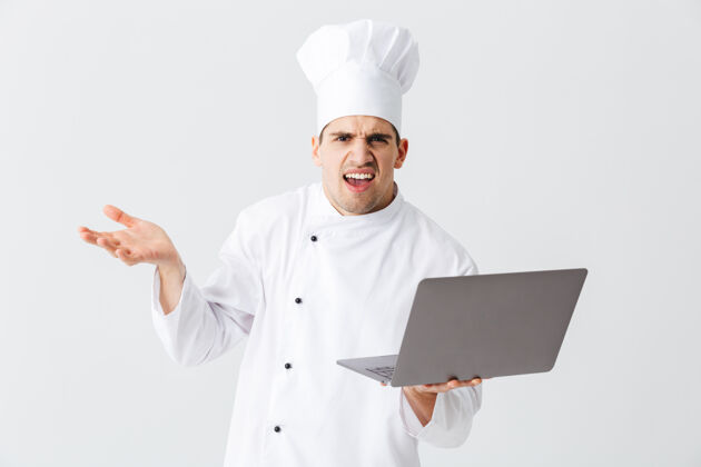 网络困惑的厨师穿着制服站在白墙上 手里拿着笔记本电脑笔记本电脑员工制服