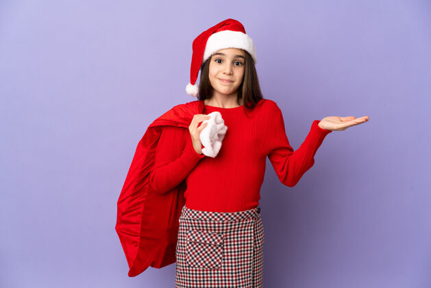 不确定戴着帽子和圣诞袋的小女孩被隔离在紫色的背景下 举手的时候有疑问怀疑圣诞老人失望