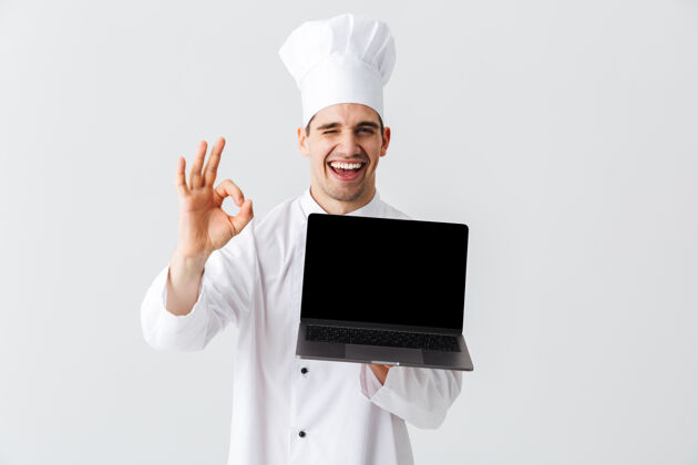 烹饪开朗的厨师身穿厨师制服站在白色的墙上 手里拿着银行屏幕的笔记本电脑职业欢呼工作