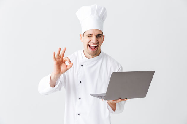 笔记本电脑开朗的厨师穿着制服站在白色的墙上 手里拿着笔记本电脑工人职业服务