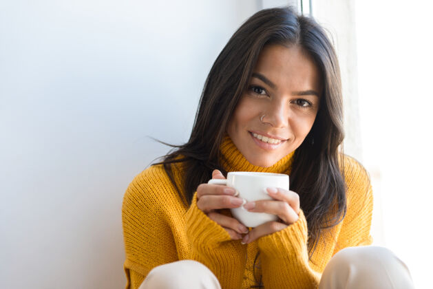 秋天特写一个微笑的年轻女子穿着毛衣坐在窗前 拿着一杯茶女舒适饮料