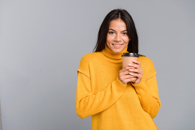 热一个微笑的年轻女子穿着毛衣 拿着外卖咖啡杯 隔着灰色的墙自信白种人人