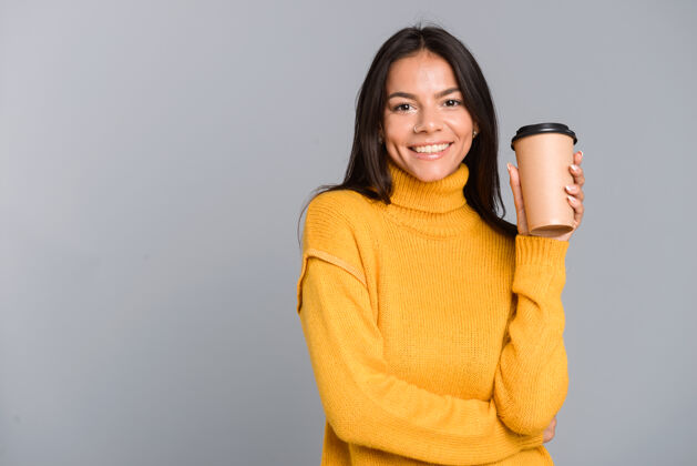 欢呼一个穿着毛衣 拿着外卖咖啡杯 隔着灰墙的快乐年轻女子的画像凉爽手咖啡