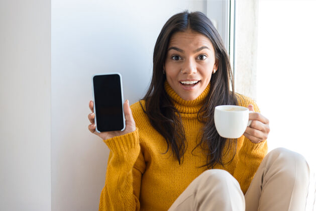 放松特写镜头中一位身着毛衣的快乐年轻女子坐在室内的窗前 捧着一杯茶 展示着一部黑屏手机微笑欢呼午餐
