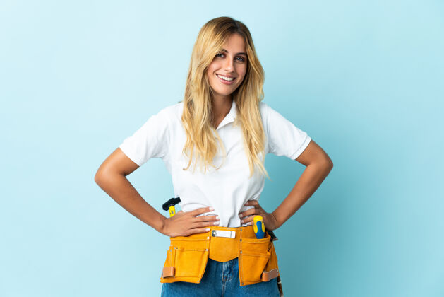 工作年轻的金发乌拉圭电工女子蓝色摆出双臂在臀部和微笑手工车间成年人