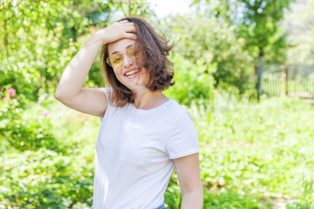 成人美丽的年轻黑发女子 棕色头发 戴着时髦的黄色太阳镜 在公园或花园的绿色背景下休息和微笑魅力新鲜笑