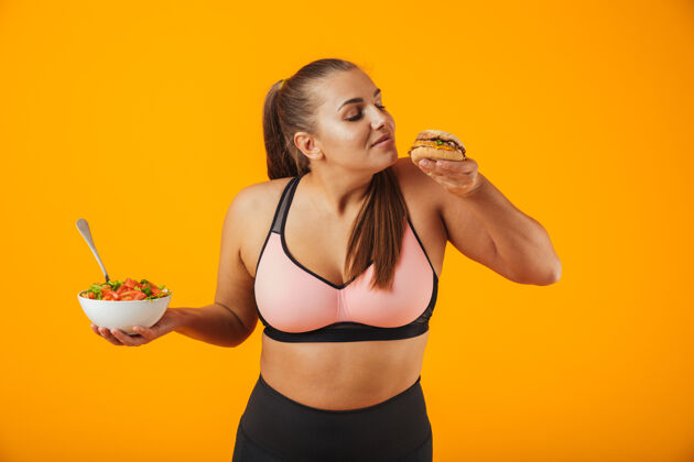 体重一个穿着运动服的快乐的超重健身妇女的肖像 孤立地站在黄色的墙上 手里拿着盛着沙拉和汉堡的碗饮食运动装食物