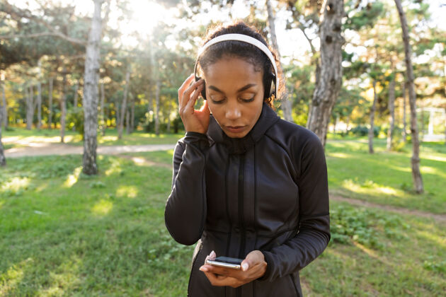 伸展图为20多岁的运动型女子穿着黑色运动服 戴着耳机 在绿色公园里散步时使用手机训练散步吸引力