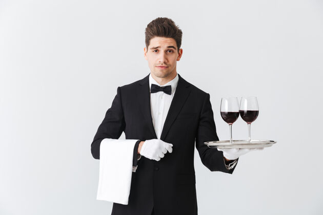 鞠躬穿着燕尾服的年轻帅哥侍者端着一个盘子 灰色的墙上隔着两杯红酒酒杯介绍杯子
