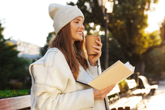 女人一个红发女人坐在户外的长凳上拿着一本书喝着咖啡咖啡坐着成人