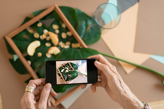 人手创意成熟女性的手拿着智能手机 在绿叶上苹果片组成的木架上拍照电脑无线技术室内