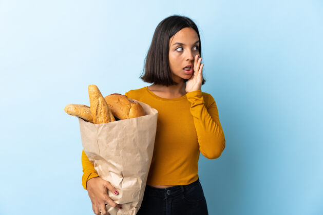 说话一个年轻的拉丁女人正在买一些孤立在蓝色地板上的面包 一边看一边用惊奇的手势低声说着什么面包对话女人