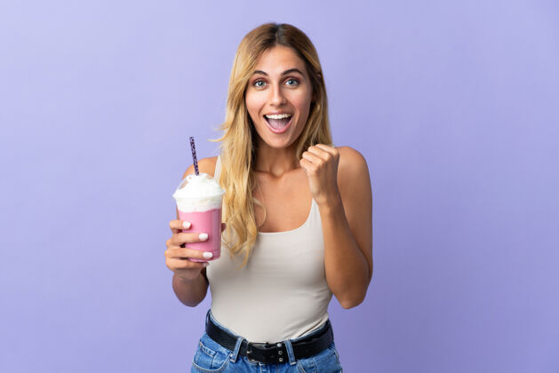 甜点年轻的金发乌拉圭妇女用草莓奶昔孤立在紫色庆祝胜利的位置年轻人提神奶昔