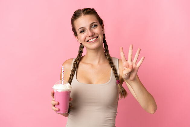成人年轻女子用草莓奶昔孤立在粉红色的快乐和计数四个手指刷新人成功