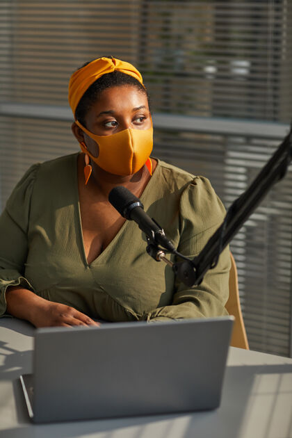 工作戴着防护面具的年轻女面试官在广播中使用笔记本电脑和麦克风讲话办公室办公桌主持