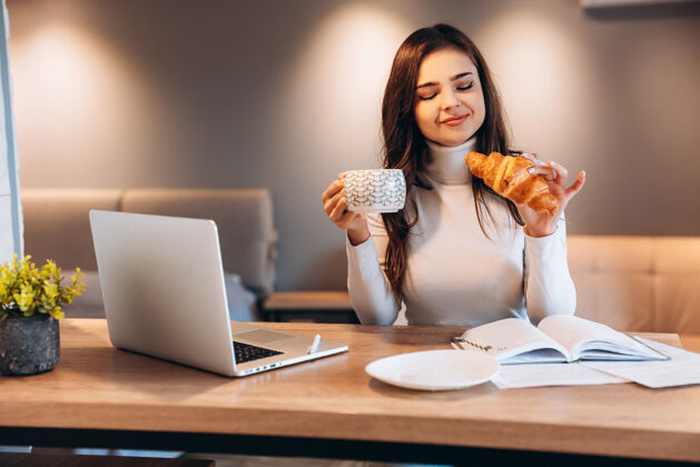 食物坐在办公室里使用笔记本电脑的自由职业女性家年轻坐在厨房里干活的女人笔记本电脑漂亮工作时喝咖啡喝茶的女性房子享受商务女性