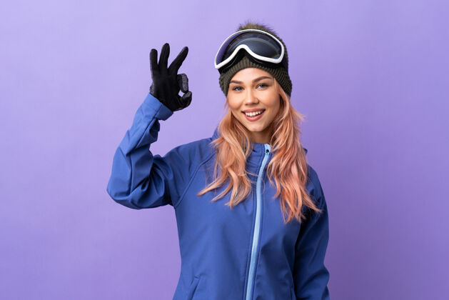 滑雪滑雪者女孩戴着滑雪板眼镜在孤立的紫色上用手指显示ok标志年轻女人玻璃