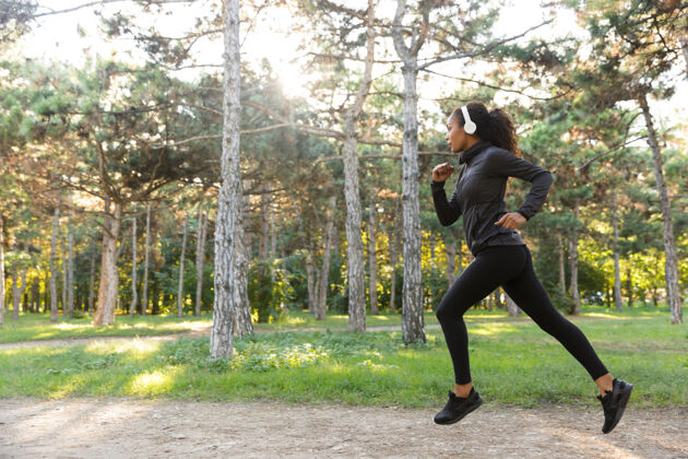 吸引力一张20多岁的女运动员穿着黑色运动服 戴着耳机 在绿色公园里跑步的照片女性耳机保健