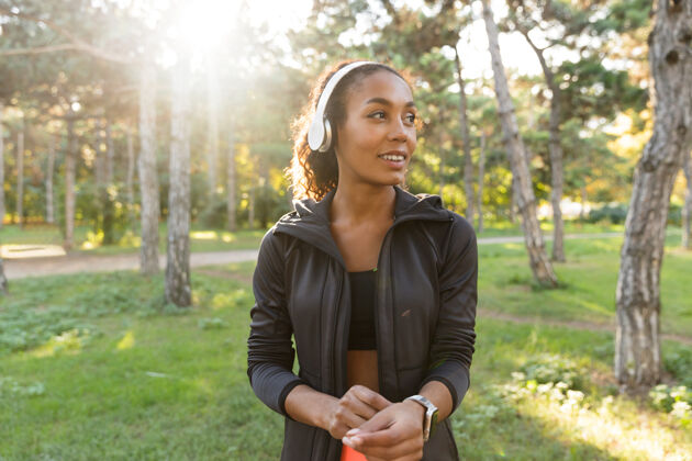 自然一个20多岁的女人穿着黑色运动服 戴着耳机 微笑着穿过绿色公园温暖公园早晨
