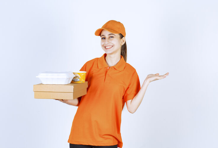 购物身着橙色制服的女快递员手里拿着一个纸板箱 一个塑料外卖箱和一个黄色的面条杯姿势年轻人员工