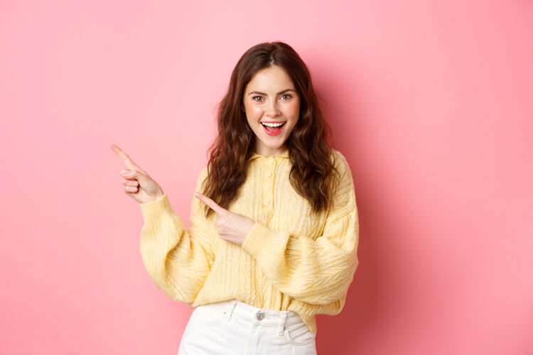 休闲自信快乐的女人 卷发 手指放在一边 在左边的版面上展示宣传片 兴奋地微笑着 站在粉色的墙上指向魅力女性