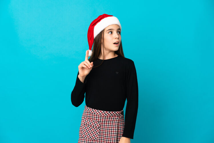 奇迹一个戴着圣诞帽的小女孩孤零零地站在蓝色的墙上 想着一个向上指指点点的主意诺埃尔节日问题