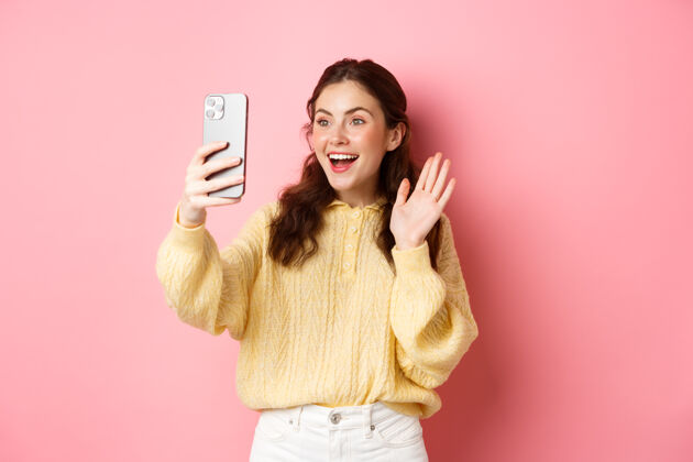 乐观开朗的女孩在智能手机上视频聊天 对着手机摄像头挥手 开心地笑着 打声招呼 站在粉色的墙上在线请应用程序