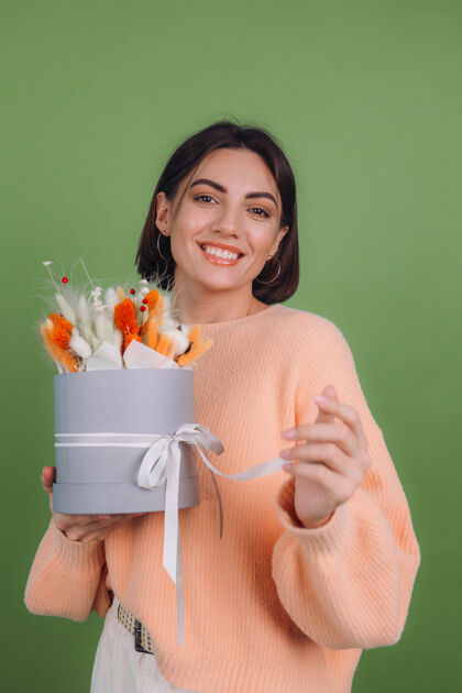 冷静年轻女子穿着休闲桃色毛衣隔离在绿橄榄墙上手持橙白色花盒组成的棉花 吉普赛拉小麦和拉古鲁斯作为礼物开心惊喜干花花束帽盒花