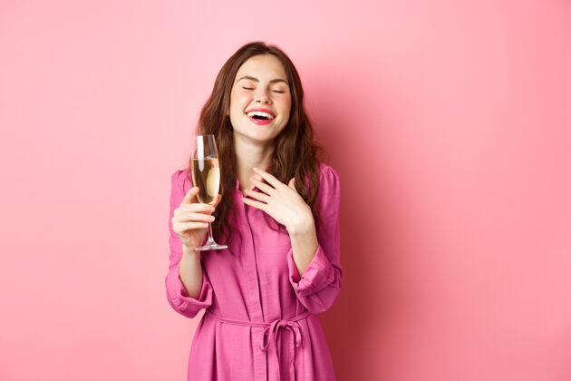 香槟庆祝和节日的概念时尚风骚的女人穿着时髦的衣服 在聚会上拿着一杯香槟 微笑着高兴 站在粉红色的墙上干杯情绪微笑