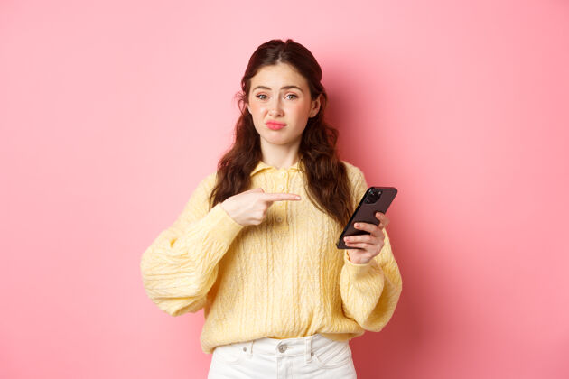女人怀疑的年轻女子不满意地做鬼脸 手指着智能手机屏幕 对网络内容有怀疑 站在粉红色的墙上应用程序手机魅力