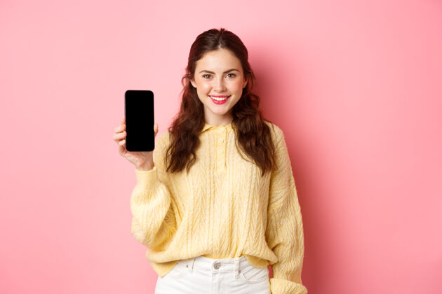 互联网科技和网上购物年轻的微笑的女人看起来坚定 建议下载应用程序 在屏幕上显示智能手机应用程序 站在粉红色的墙壁上水平情绪黑发