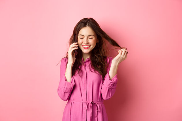 女性美丽的年轻女子打电话给别人 在打电话的时候笑嘻嘻的 和朋友聊天 玩发丝 站在粉红色的墙上购物乐观应用程序