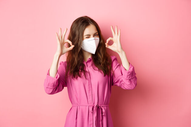 呼吸器Covid-19 流行性疾病和生活方式概念开朗的女孩表现出良好的迹象 戴医用呼吸器作为预防电晕的措施 粉红色的墙壁复制空间女孩流行请