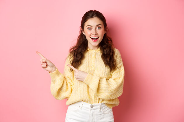 学生年轻兴奋的女人穿着休闲装 展示横幅 手指着左边的复制空间 站在粉红色的墙上惊人请时尚