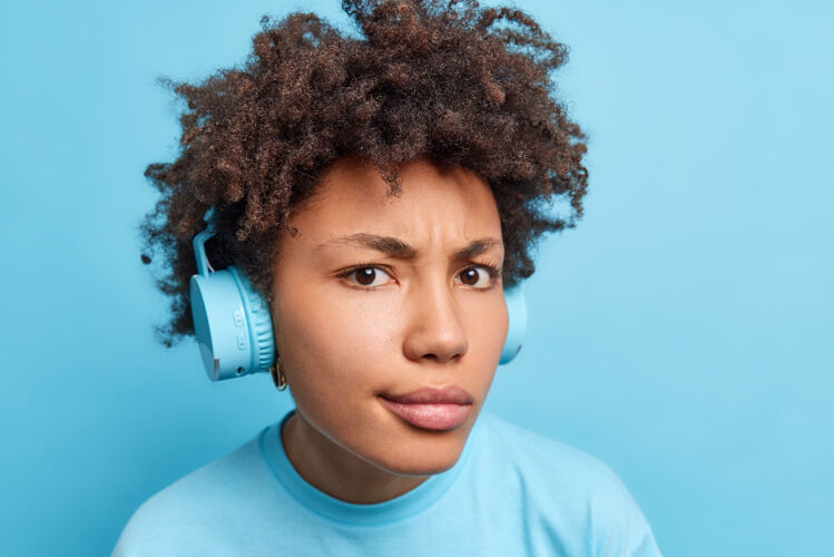 歌曲严重不高兴的卷发非洲裔美国妇女戴立体声无线耳机看着专心听音乐或有声书穿着随意隔离在蓝色的墙壁肖像民族女人