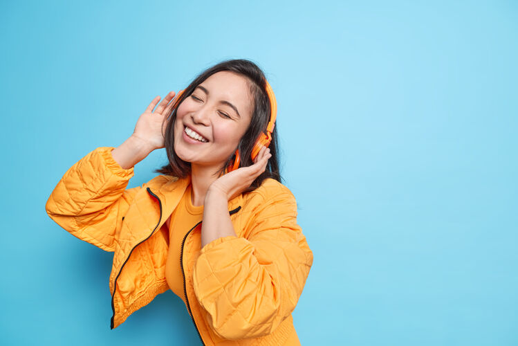 微笑年轻可爱的亚洲女人睁大眼睛 微笑 表情愉快 戴着耳机听音乐 穿着时髦的衣服 在蓝色的墙上欣赏最喜欢的歌曲可爱时尚人