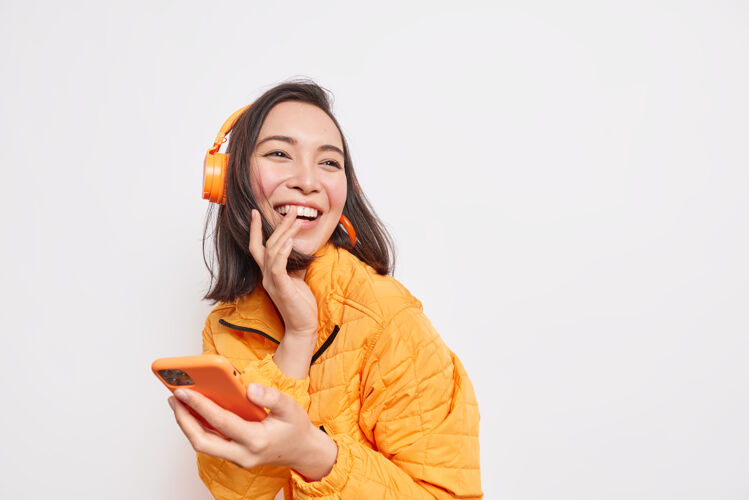 模特快乐的亚洲女人欢笑快乐地看着远方享受最喜爱的音乐播放列表使用移动应用程序戴着无线耳机穿着橙色夹克隔离在白色墙壁空白处积极女性黑发