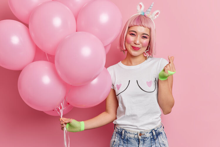 欢呼快乐的粉红色头发的女人的肖像与东方外观手势迷你标志手势姿势与氦气球享受节日穿着时髦的衣服快乐美丽微笑