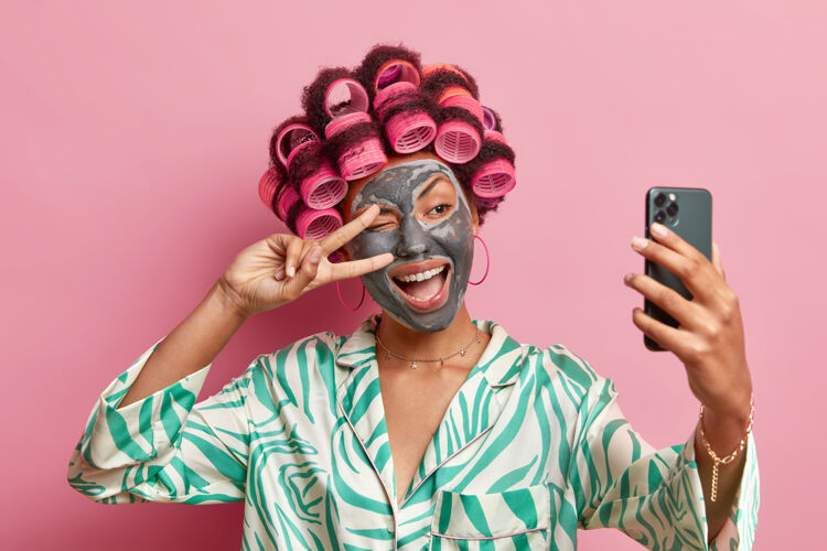 姿态高兴的少数民族妇女的照片使和平的姿态在眼睛微笑广泛应用美容面具头发辊在手机上自拍肖像穿着晨衣隔离在粉红色的墙上宠爱关怀黏土