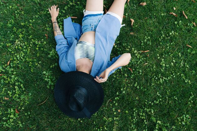 模特穿着蓝色条纹夹克和黑色夹克的年轻女子的时尚肖像帽子女人躺在公园的绿色草坪上公园孤独姿势